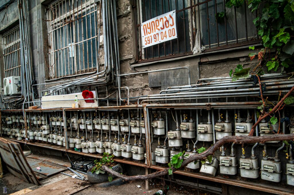 Row of outdoor gas meters on a building facade with vines and a sign in non-Latin script.